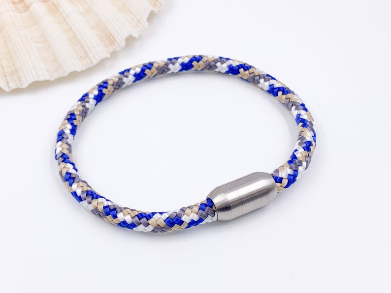 Puede incluir: Un brazalete de cuerda trenzada azul, blanco y beige con un cierre magn&eacute;tico plateado.