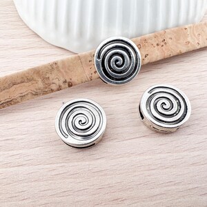 Pode incluir: Três contas redondas de metal prateadas com um design em espiral. As contas estão sobre uma superfície de cortiça.