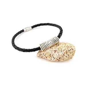 Pode incluir: Uma pulseira de couro trançado preto com um fecho cilíndrico prateado com um design em espiral. A pulseira é exibida em uma pedra clara e texturizada contra um fundo branco.