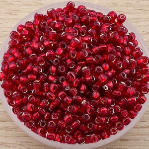 Pode incluir: Uma tigela cheia de contas de sementes de vidro vermelho. As contas têm um brilho metálico e são perfeitas para fazer joias.