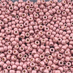 May include: A close-up image of a large quantity of small, round, pink beads. The beads have a matte finish and a silver-colored hole in the center.