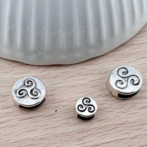 Op de afbeelding: Drie zilveren metalen bedels met een Keltisch knoopontwerp. De bedels zijn rond en hebben een gat om aan een armband of ketting te bevestigen.