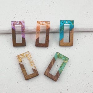 Pode incluir: Cinco pingentes retangulares com moldura de madeira e preenchimento de resina. A resina é preenchida com flocos de ouro e vem em várias cores: roxo, laranja, turquesa e verde. Os pingentes são para fazer joias.