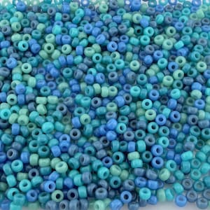 Puede incluir: Primer plano de un montón de pequeñas cuentas redondas de semillas en varios tonos de azul y verde. Las cuentas están dispersas, creando una superficie texturizada. Los colores varían del turquesa claro al azul marino oscuro.