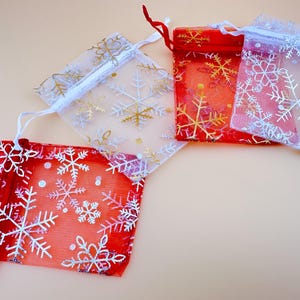 Könnte beinhalten: Vier kleine, transparente Geschenkbeutel in Rot und Weiß, verziert mit Schneeflockenmustern. Die Beutel haben Kordelzüge und sind auf einer hellbeige Oberfläche angeordnet. Einige Schneeflocken sind goldfarben, andere silbern.