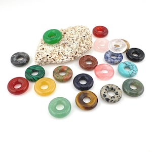 May include: A selection of gemstone donut pendants in various colours, including green, red, yellow, and blue. Some pendants have unique patterns and textures. A piece of textured stone is also present.