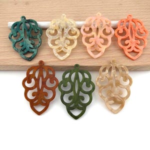 Op de afbeelding: Zeven decoratieve hangers in diverse kleuren, waaronder teal, beige, perzik en bruin. Elke hanger heeft een open ontwerp en een lus om op te hangen. De hangers liggen op een houten oppervlak.