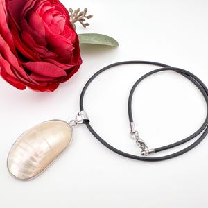 Könnte beinhalten: Ein weißer Perlmutt-Anhänger mit einer schwarzen Schnurkette. Der Anhänger ist oval geformt und hat einen natürlichen irisierenden Glanz.