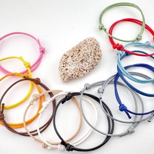 May include: A collection of adjustable bracelets in various colors, including pink, yellow, orange, brown, white, black, gray, blue, red, and green. Each bracelet features a knotted design and is made of a cord material. A textured, light-colored stone is also present.