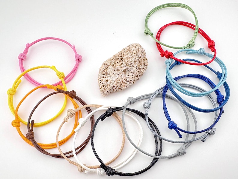 May include: A collection of adjustable bracelets in various colors, including pink, yellow, orange, brown, white, black, gray, blue, red, and green. Each bracelet features a knotted design and is made of a cord material. A textured, light-colored stone is also present.