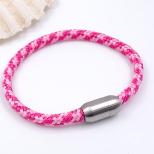 Puede incluir: Un brazalete de cuerda trenzada rosa y blanca con un cierre magn&eacute;tico plateado.