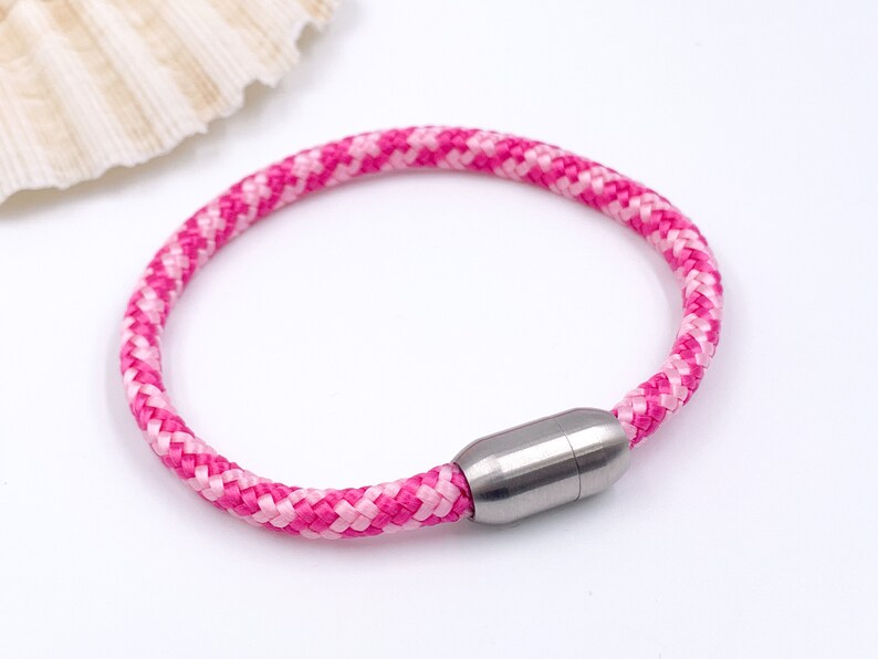 Puede incluir: Un brazalete de cuerda trenzada rosa y blanca con un cierre magn&eacute;tico plateado.