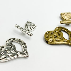 Op de afbeelding: Twee hartvormige metalen sluitingen met een getextureerd oppervlak. Eén sluiting is zilver en de andere is goud.