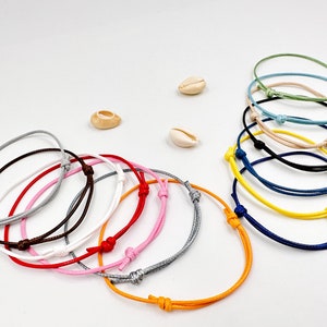 May include: A collection of adjustable cord bracelets in various colors, including gray, pink, red, brown, white, and orange. Several bracelets are stacked together, with additional colors like blue, yellow, and green. Small seashells are scattered around.
