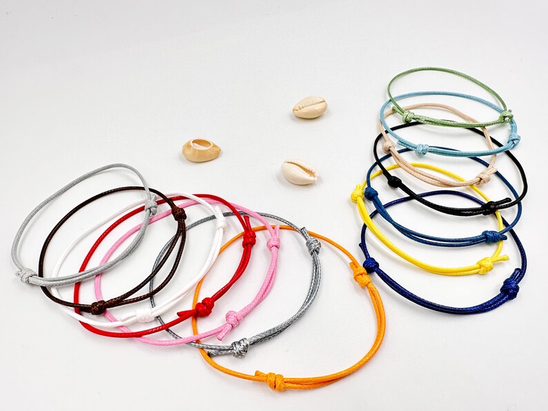 May include: A collection of adjustable cord bracelets in various colors, including gray, pink, red, brown, white, and orange. Several bracelets are stacked together, with additional colors like blue, yellow, and green. Small seashells are scattered around.
