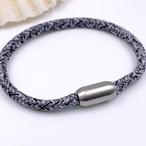 Puede incluir: Un brazalete trenzado gris con un cierre magn&eacute;tico plateado.