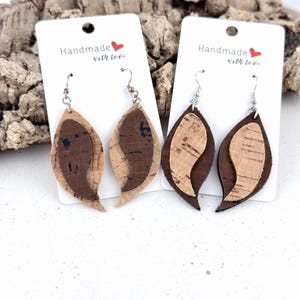 Pode incluir: Dois pares de brincos pendentes em forma de folha feitos de material de cortiça em camadas. O primeiro par apresenta cortiça castanho-clara com inserções castanho-escuras, enquanto o segundo tem cortiça castanho-escura com inserções castanho-claras.