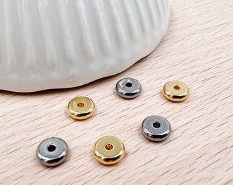 Puede incluir: Seis pequeñas cuentas redondas de metal en tonos dorados y plateados. Cada cuenta tiene un agujero en el centro. Las cuentas están dispersas sobre una superficie de madera clara, con un plato de cerámica blanco en el fondo.