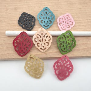 Op de afbeelding: Een verzameling van acht decoratieve filigraan hangers in verschillende kleuren, waaronder zwart, groenblauw, lichtroze, rood, beige, groen, geel en donkerroze. Elke hanger heeft een ingewikkeld, symmetrisch uitgesneden patroon en een gaatje.