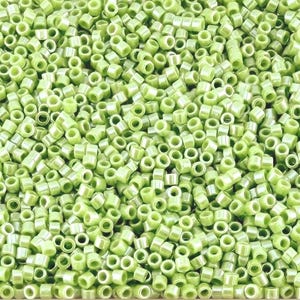 May include: Close-up of a pile of light green seed beads. The beads are cylindrical with a hole running through the center. The beads are small and uniform in size, creating a textured surface.