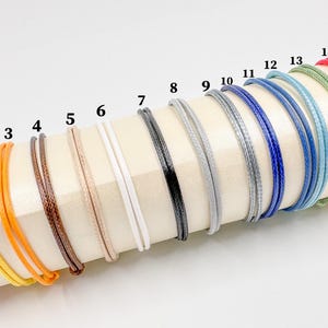 May include: A collection of colorful bracelets in various shades, including pink, yellow, orange, brown, beige, white, black, gray, blue, and green. Each bracelet is made of a thin, woven cord, and they are displayed on a cream-colored surface.