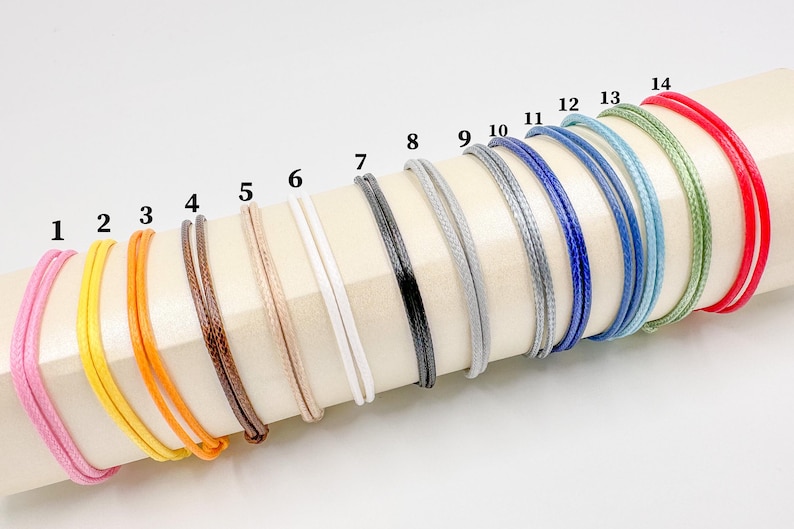 May include: A collection of colorful bracelets in various shades, including pink, yellow, orange, brown, beige, white, black, gray, blue, and green. Each bracelet is made of a thin, woven cord, and they are displayed on a cream-colored surface.