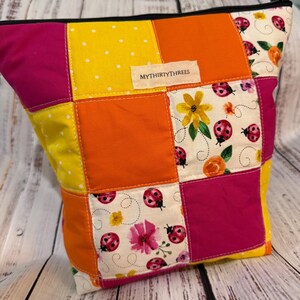 May include: A patchwork zippered pouch with a vibrant mix of colors. The design includes squares of yellow with white polka dots, orange, and hot pink. Some squares feature a floral and ladybug print. The label reads "MYTHIRTYTHREE".