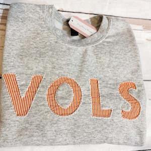 May include: A gray sweatshirt with the word "VOLS" in orange and white striped letters. The sweatshirt has a crew neck and a tag attached. The background is a light-colored wooden surface.