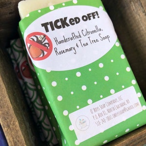 May include: A green and white bar of soap wrapped in a green paper with white polka dots. The paper has a white oval sticker with the text "TICKED OFF! Handcrafted Citronella, Rosemary & Tea Tree Soap" and a cartoon tick with a red circle and a diagonal line through it. The bottom of the paper has a white sticker with the text "O' BOYS SOAP COMPANY, LLC P.O. BOX 81, NORTH CHATHAM, NY 518-248-3807: OBOYSSOAP@GMAIL.COM".