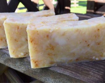 May include: Three bars of handmade soap, each with a light yellow color and a slightly grainy texture. The soap bars are arranged in a row on a wooden surface.