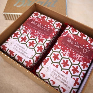 May include: Two bars of handcrafted scented soap, wrapped in paper with a red and white floral pattern, are presented in a brown box. The soap packaging features the text "Be Berry!" and the company's name and contact information.