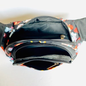 Puede incluir: Una ri&ntilde;onera negra con un estampado colorido de personajes de dibujos animados. La ri&ntilde;onera tiene dos compartimentos con cremallera y un bolsillo con cremallera en la parte delantera.