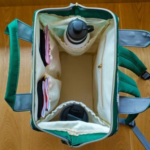 May include: A green and gray backpack with a white interior. The backpack is open and has several pockets and compartments. There are items inside the backpack, including a water bottle, a cup, and some small items.