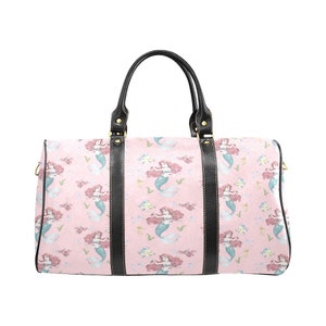 May include: A pink duffel bag featuring a repeating pattern of mermaids with red hair and blue tails. The bag has black leather handles and trim. The bag is designed for travel.