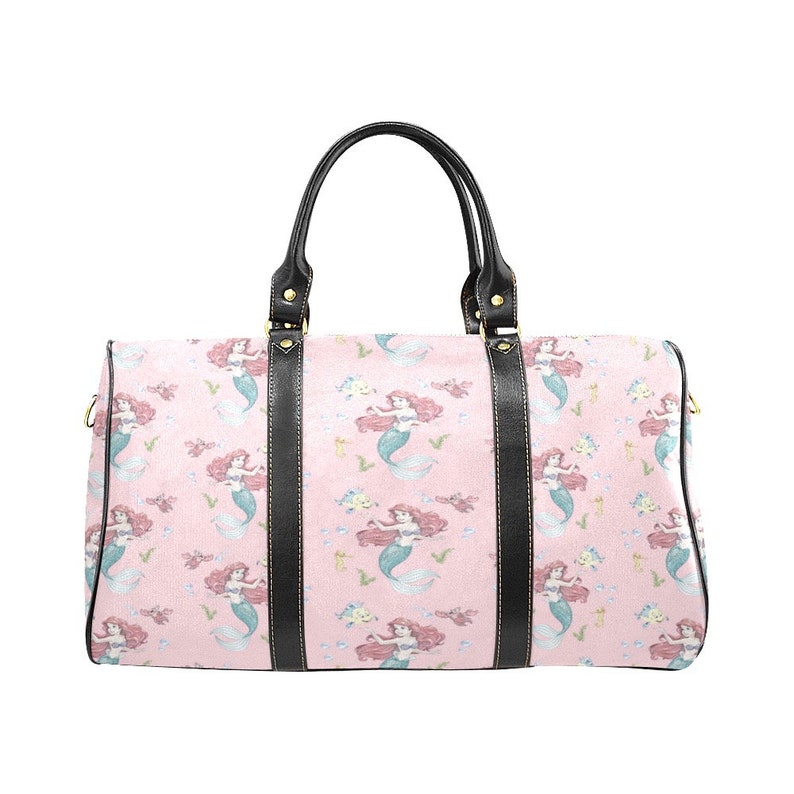 May include: A pink duffel bag featuring a repeating pattern of mermaids with red hair and blue tails. The bag has black leather handles and trim. The bag is designed for travel.