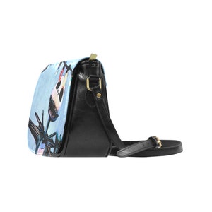 Puede incluir: Un bolso bandolera de cuero negro con un dise&ntilde;o estampado en azul y blanco que presenta un personaje de dibujos animados con una cara de calavera. El bolso tiene una hebilla dorada y una correa larga ajustable.