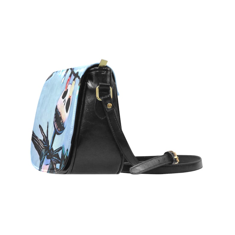 Puede incluir: Un bolso bandolera de cuero negro con un dise&ntilde;o estampado en azul y blanco que presenta un personaje de dibujos animados con una cara de calavera. El bolso tiene una hebilla dorada y una correa larga ajustable.