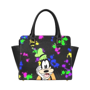 Puede incluir: Bolso de mano negro con un diseño de salpicaduras de colores y un personaje de dibujos animados. El bolso tiene un asa superior y bolsillos laterales. El personaje de dibujos animados lleva un sombrero verde y una camisa naranja.