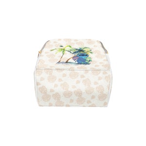 Puede incluir: Una bolsa de cosm&eacute;ticos blanca con un patr&oacute;n de hojas de palma marr&oacute;n claro. La bolsa presenta un personaje azul Stitch de Lilo & Stitch sosteniendo una tabla de surf verde. La bolsa tiene una cremallera dorada.