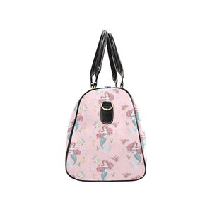 May include: A pink duffel bag featuring a repeating pattern of mermaids with red hair and teal tails. The bag has black handles and trim, with a gold-colored buckle detail. The bag is designed for travel or carrying items.