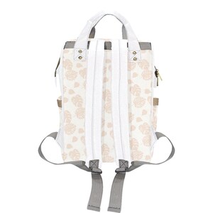 Puede incluir: Una mochila de lona blanca con un estampado floral rosa. La mochila tiene correas ajustables y un asa superior.