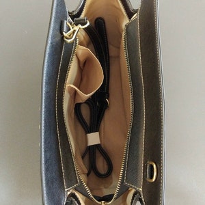 K&ouml;nnte beinhalten: Schwarze Handtasche, von oben gesehen, mit offenem Innenraum, beigem Futter und mehreren F&auml;chern. Die Tasche hat einen goldenen Rei&szlig;verschluss, einen schwarzen Riemen und goldfarbene Beschl&auml;ge. Die Au&szlig;enseite ist schwarz mit wei&szlig;en N&auml;hten.