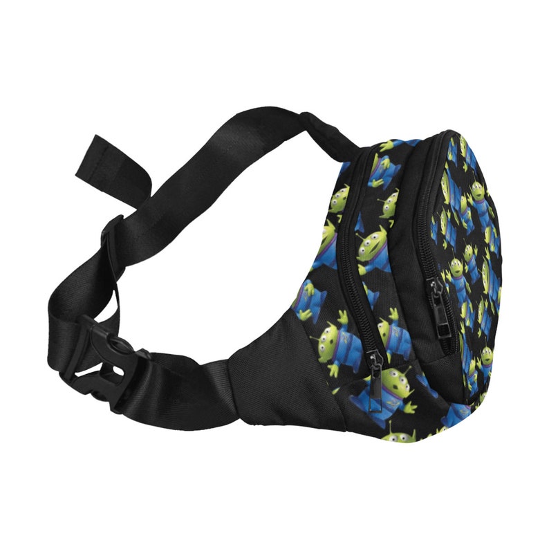 Puede incluir: Una ri&ntilde;onera negra con un patr&oacute;n repetido de extraterrestres verdes con ropa azul. La ri&ntilde;onera tiene dos compartimentos con cremallera y una correa negra ajustable.