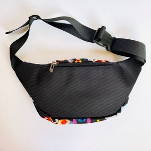 Puede incluir: Ri&ntilde;onera negra con un estampado colorido de personajes de dibujos animados. La ri&ntilde;onera tiene un cierre de cremallera y una correa ajustable con hebilla.