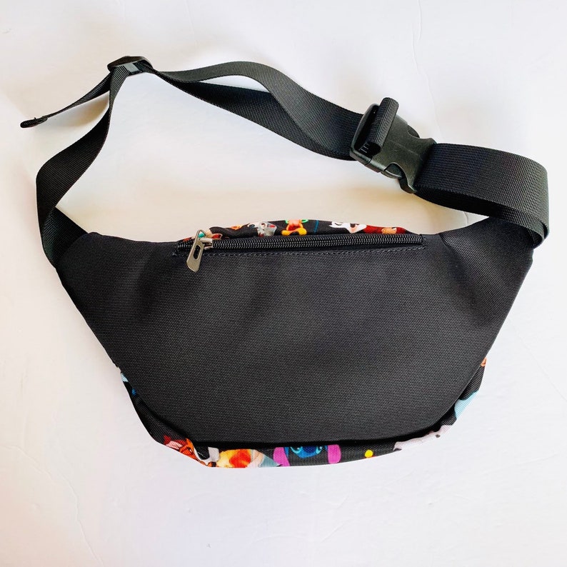 Puede incluir: Ri&ntilde;onera negra con un estampado colorido de personajes de dibujos animados. La ri&ntilde;onera tiene un cierre de cremallera y una correa ajustable con hebilla.