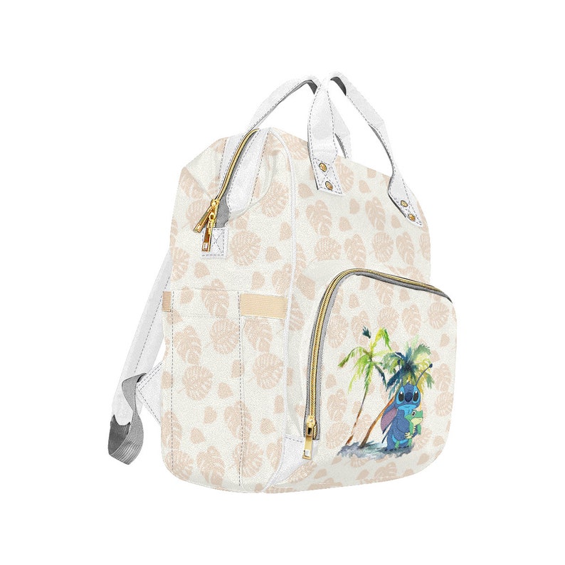 Puede incluir: Bolso de pa&ntilde;ales blanco con un estampado de hojas tropicales y un personaje azul Stitch de Lilo & Stitch en la parte delantera. El bolso tiene una cremallera dorada y un asa dorada.