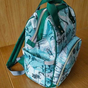 May include: A green and blue patterned backpack with a large front pocket and adjustable straps. The backpack has a zipper closure and a gold zipper pull.