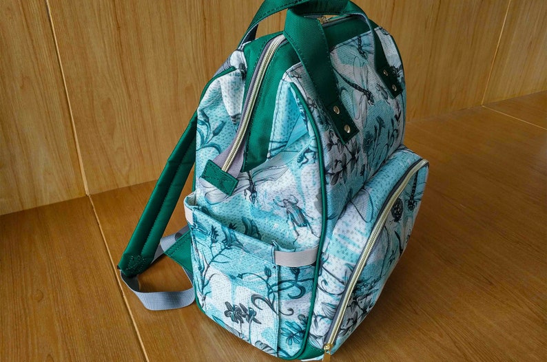 May include: A green and blue patterned backpack with a large front pocket and adjustable straps. The backpack has a zipper closure and a gold zipper pull.