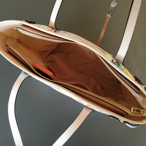 Pu&ograve; includere: Una borsa tote bianca con stampa floreale e una cerniera dorata. La borsa ha due manici bianchi e una tasca con cerniera all'interno.