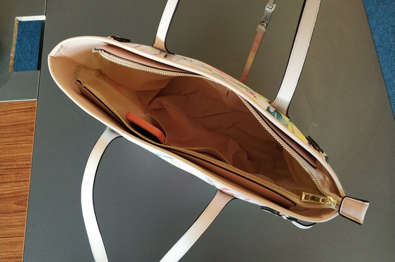 Pu&ograve; includere: Una borsa tote bianca con stampa floreale e una cerniera dorata. La borsa ha due manici bianchi e una tasca con cerniera all'interno.